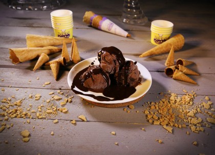 lody czekoladowe, podane na białym, porcelanowym talerzyku, polane roztopioną czekoladą, wokół waflowe rożki i papierowe miseczki